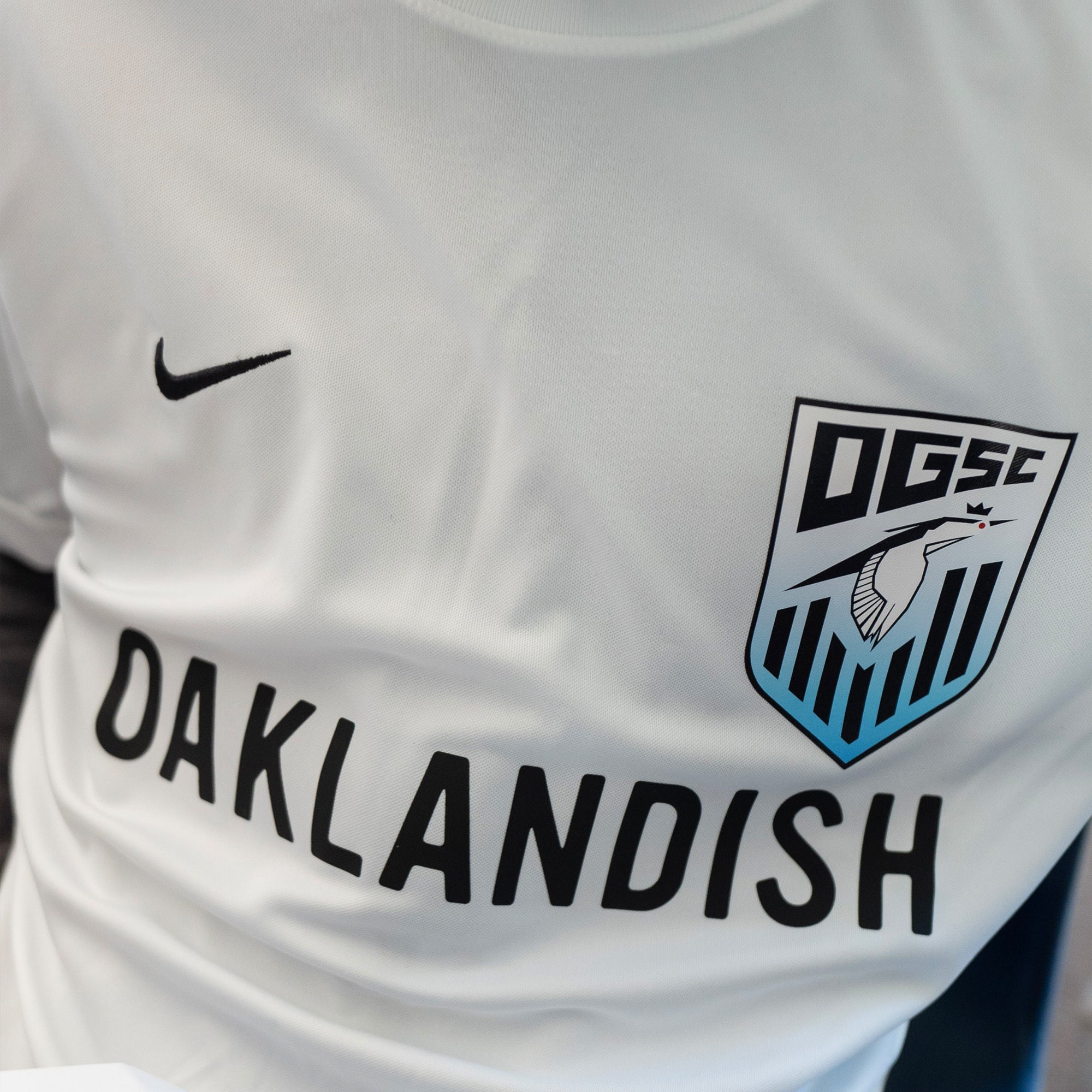Close-up of a person wearing the Oakland Genesis Home White sports jersey by Partner, featuring the Nike logo on the left chest. The jersey showcases a shield emblem with "DGSC" and an image of a bird perched on a branch, accompanied by the text "Oaklandish" below. This jersey represents Oakland Genesis, a nonprofit organization dedicated to youth soccer. The background is out of focus.