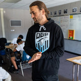 A man standing in a classroom wearing a black pullover hoodie with the Oakland Genesis soccer league heron logo on the chest.