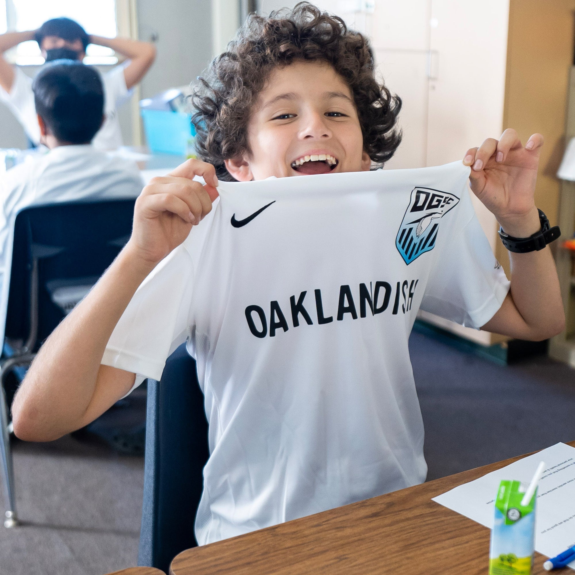 A child with curly hair and a big smile sits at a classroom desk, proudly holding up the front of an "Oakland Genesis Home White" sports jersey by Partner that reads "OAKLANDISH" and features the "DFC" logo. This Oakland Genesis jersey symbolizes their involvement in youth soccer through a nonprofit organization. A juice box rests on the desk, and another student is visible but out of focus in the background.