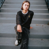 Man sitting on steps in black long sleeve tee with a gold Oaklandish tree logo, white wordmark on chest, and grey trim on neck.