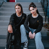 Woman and man sitting on steps in black long-sleeve tees with a gold Oaklandish tree logo, white wordmarks, and grey trim on the neck.