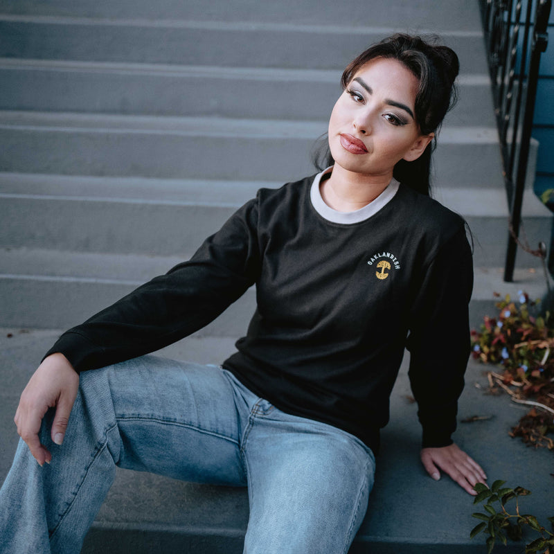 Woman sitting on steps in a black long-sleeve tee with a gold Oaklandish tree logo, white wordmark, and grey trim on the neck.