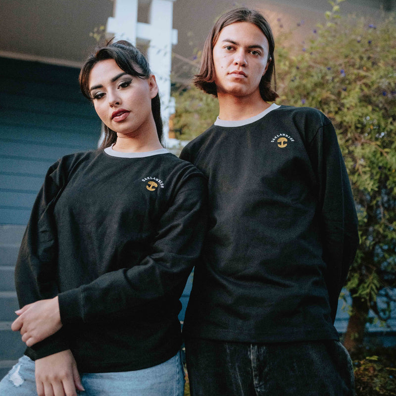 Woman and man outside in black long-sleeve tees with a gold Oaklandish tree logo, white wordmarks, and grey trim on the neck.