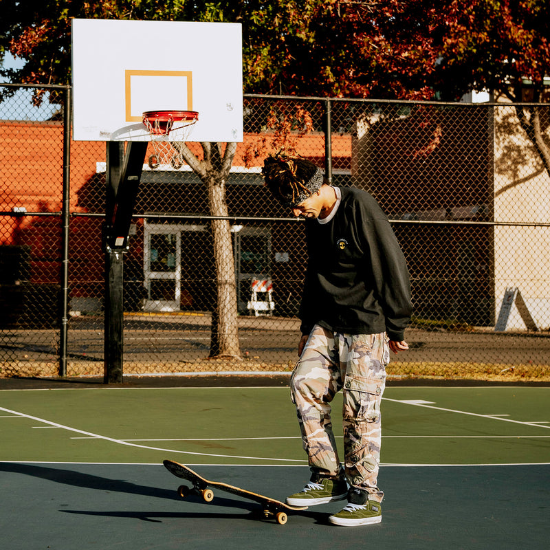 Male model skateboarding wearing black long sleeve crew neck tee with a gold Oaklandish tree logo, white wordmark on the left chest, and grey trim on neck.