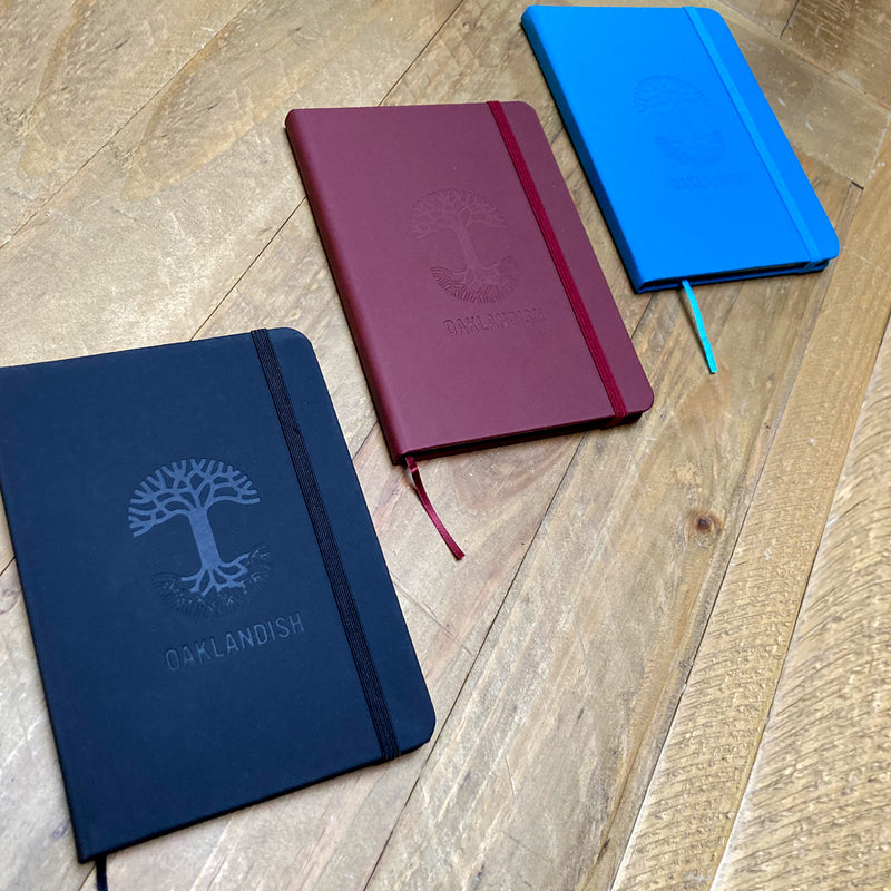  The three colorways (black, blue, and burgundy) of Oaklandish journal notebooks each debossed Oaklandish tree logo & wordmark on a wooden surface.