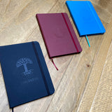 Three colorways (black, blue, and burgundy) of Oaklandish journal notebooks debossed Oaklandish tree logo & wordmark on a wood surface.