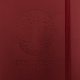 Detailed close-up of embossed Oaklandish tree logo on the front of a red journal notebook.