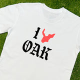 White t-shirt with ”I Oak” in black font and a red map of Oakland replacing the heart between I and Oak lying on grass.
