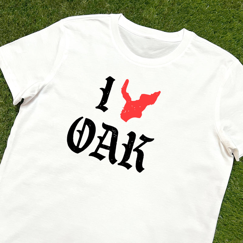 White women's t-shirt with ”I Oak” in black font and a red map of Oakland replacing the heart between I and Oak lying on the grass.