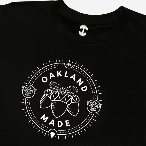 Close up of white hand-drawn Oakland Made wordmark and chesnuts in a circular graphic on chest of woman’s cut black t-shirt.