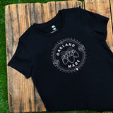 Women’s black t-shirt with white hand-drawn Oakland Made wordmark and chesnuts on chest lying out doors on wood deck on grass.