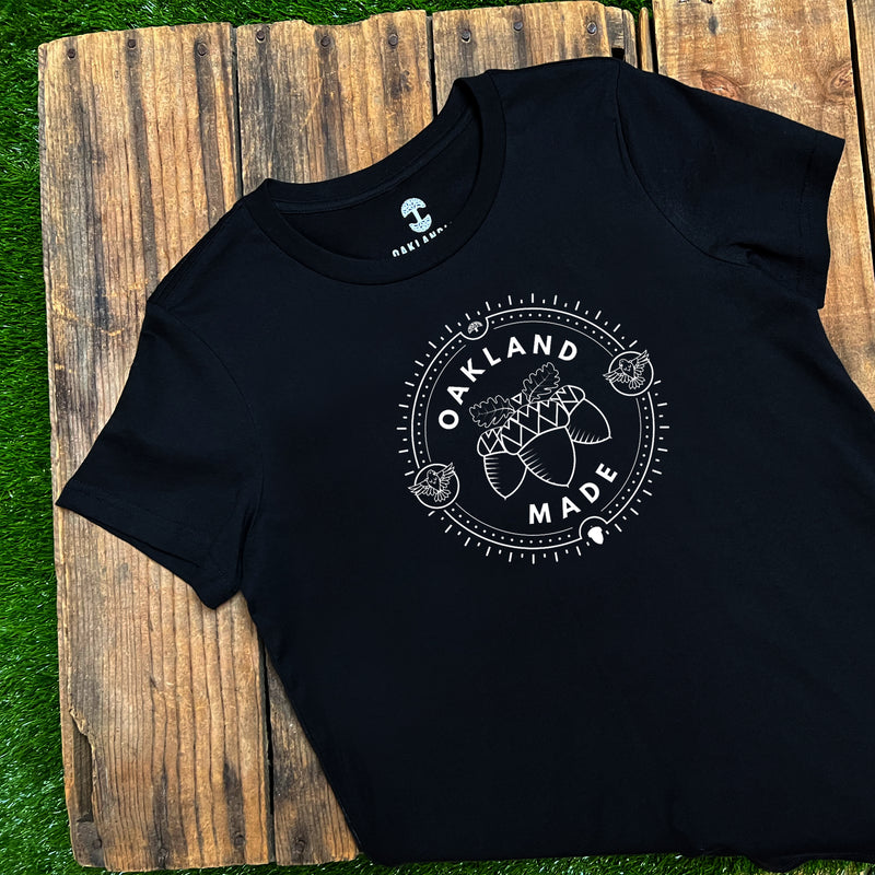 Women’s black t-shirt with white hand-drawn Oakland Made wordmark and chesnuts on chest lying out doors on wood deck on grass.