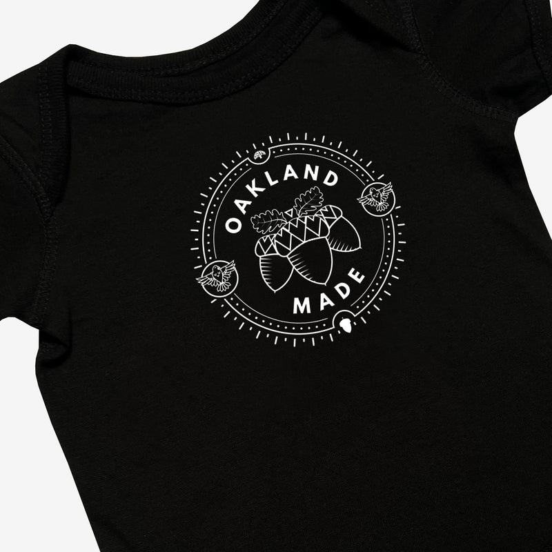 Close-up of black infant one-piece with a white circle graphic with the words “Oakland Made” and hand-drawn chestnuts. 