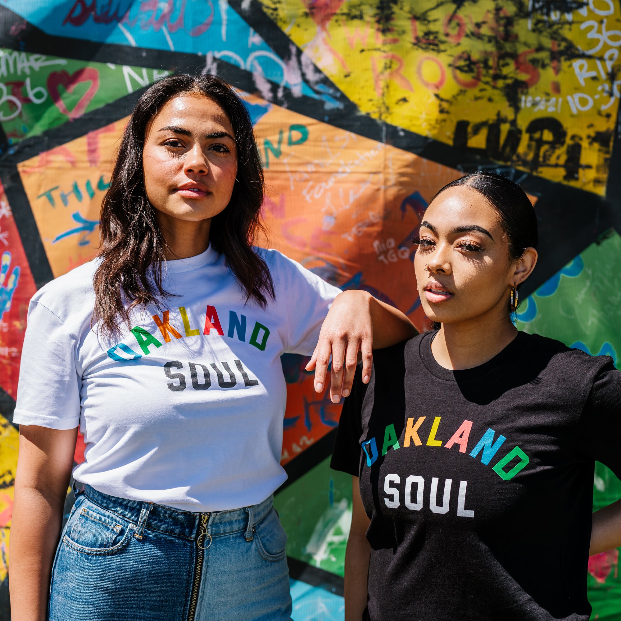 Two woman standing, with an arm resting on the other’s shoulder & graffiti background. Both wearing Oakland Soul Wordmark T-Shirts.