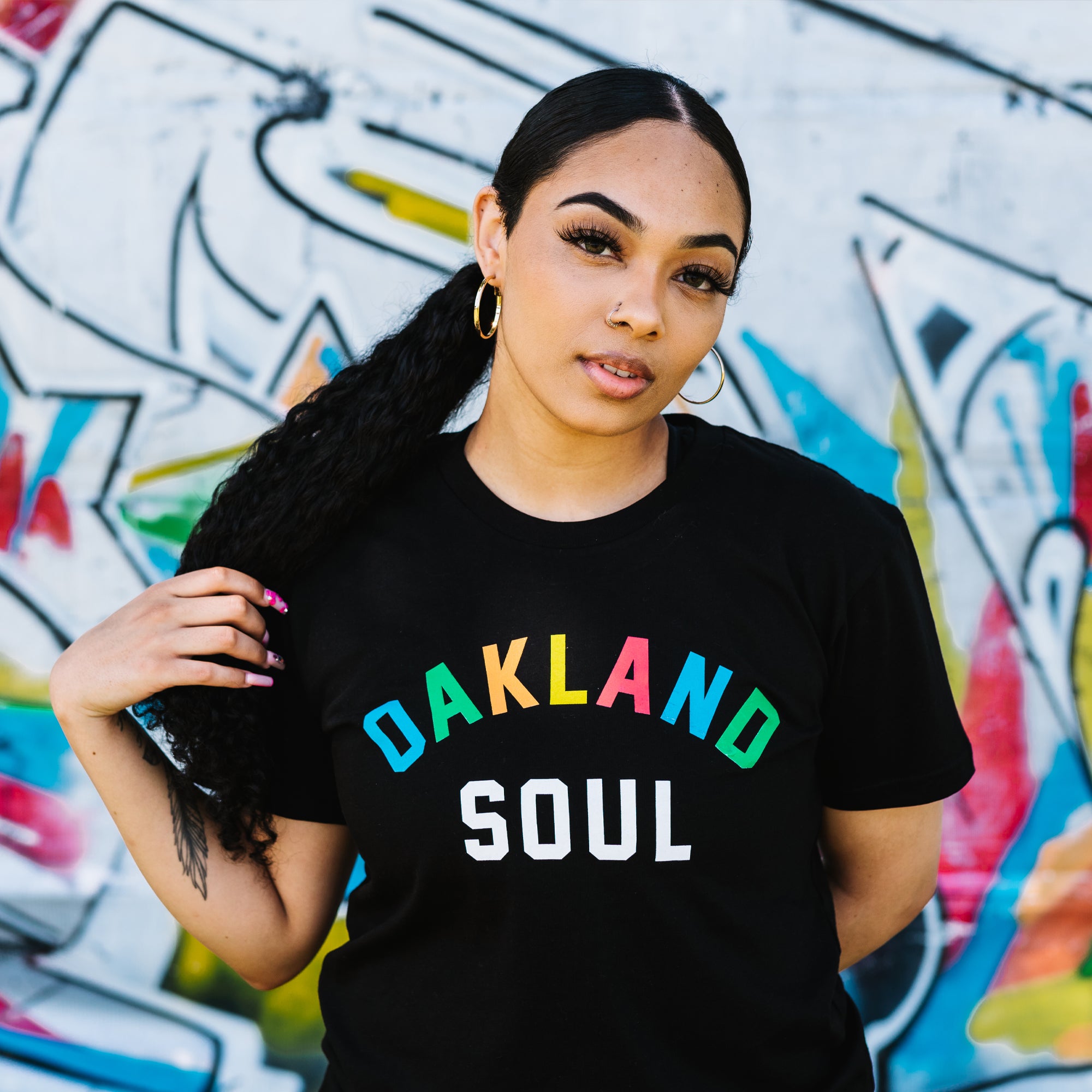 Woman from waist up, wearing black t-shirt with full color Oakland Soul wordmark logo. Blurred graffiti background.