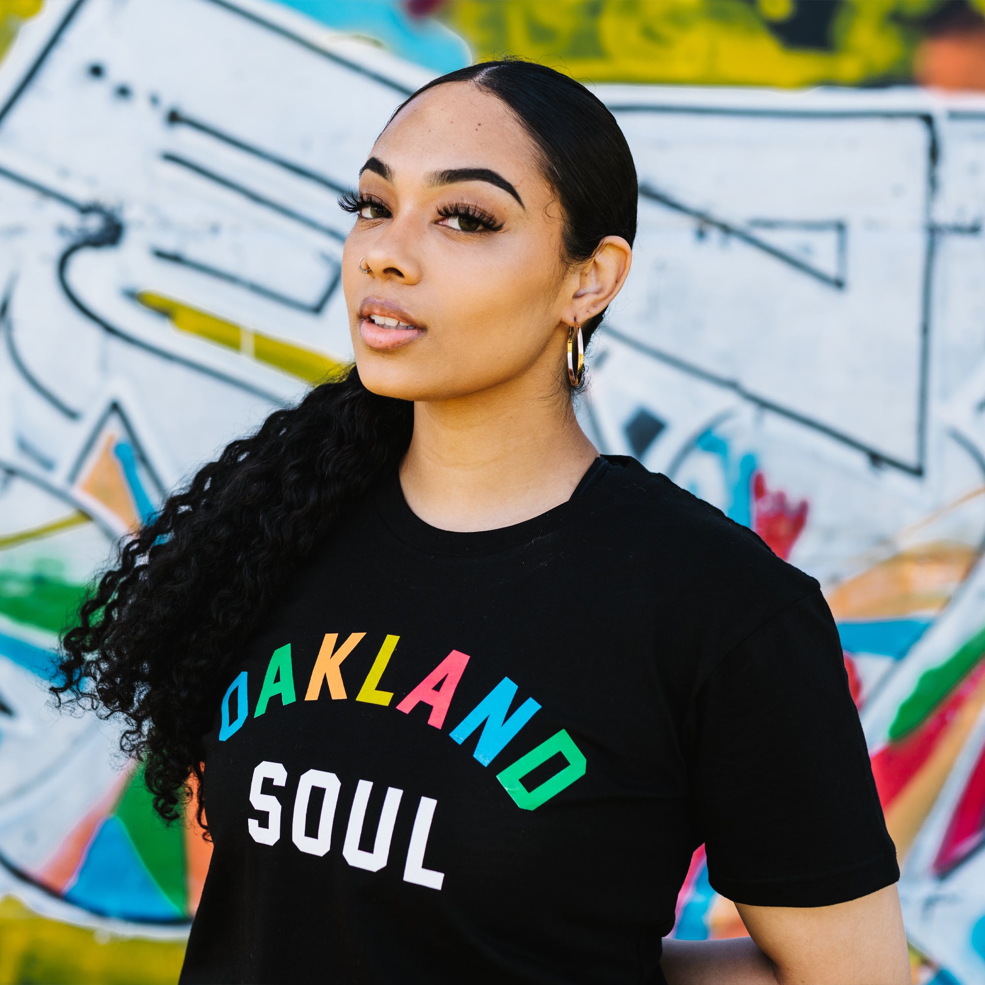 Front view of woman from waist up wearing black t-shirt with full color Oakland Soul wordmark logo. Blurred graffiti background.