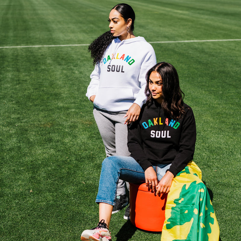 Two women outdoors on a soccer field, one wearing a black Oakland Soul hoodie and the other wearing a white one.