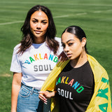 Two woman standing with waist up view, one draped in Oakland flag, soccer field background. Both wearing Oakland Soul Wordmark T-Shirts.