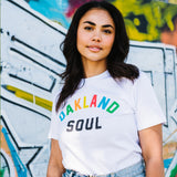Woman from waist up, wearing white t-shirt with full color Oakland Soul wordmark logo. Blurred graffiti background.