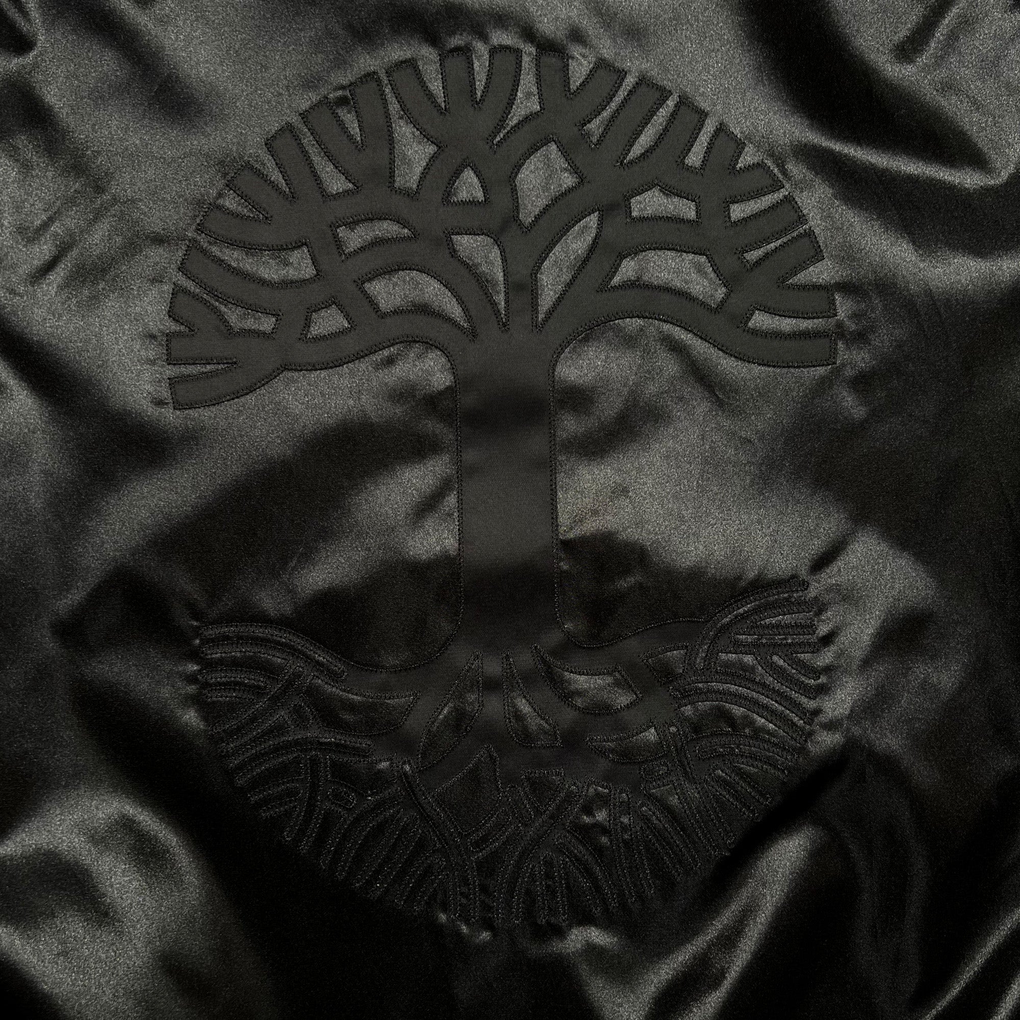 Detailed close-up of large centered Oaklandish tree logo on the backside of Mitchell & Ness black satin reversible jacket.