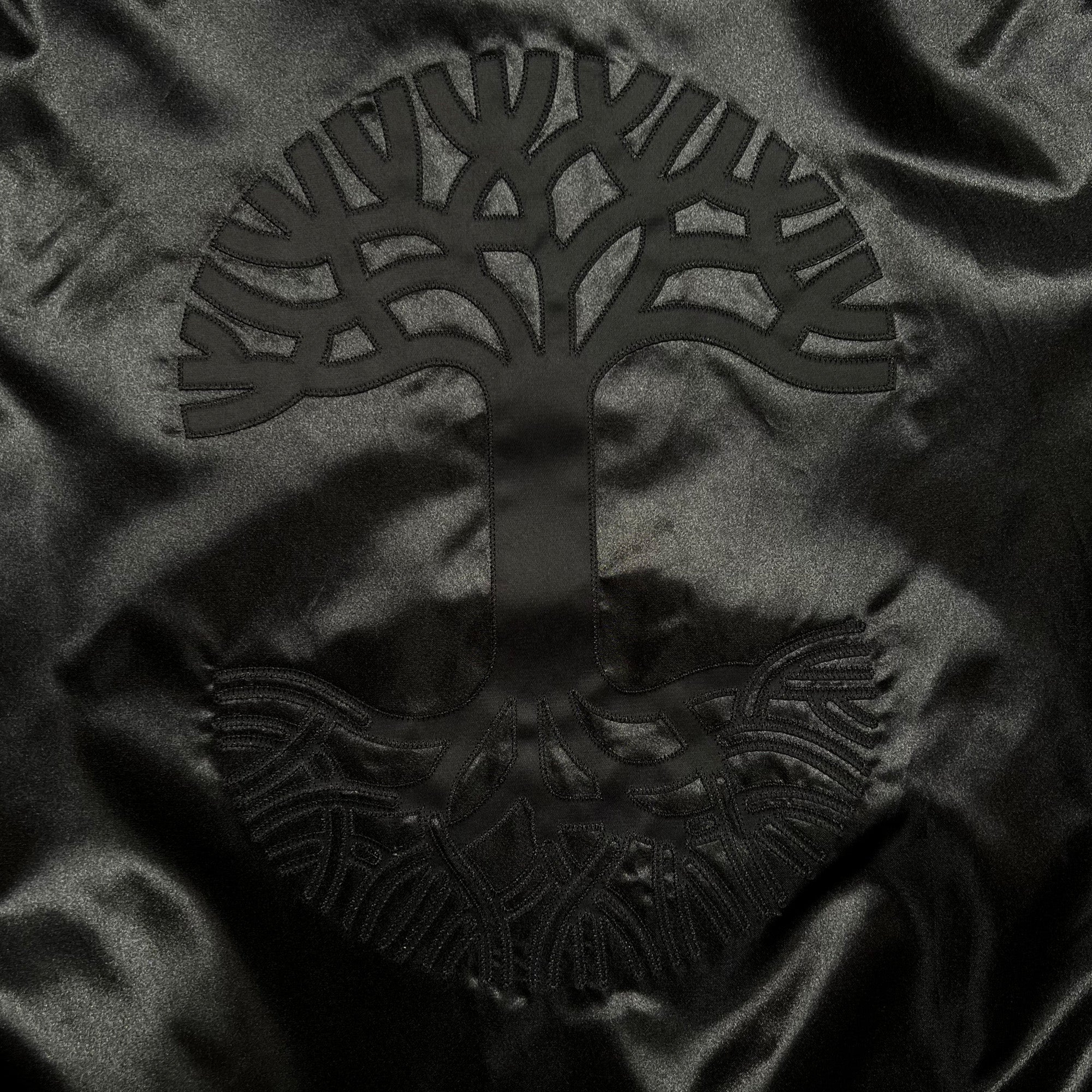The Mitchell & Ness x Oaklandish Reversible Satin Jacket features a dark green satin finish with an embroidered tree design. The symmetrical, circular motif of interwoven branches and roots echoes Oaklandish style, while the shiny texture enhances its monochrome depth.