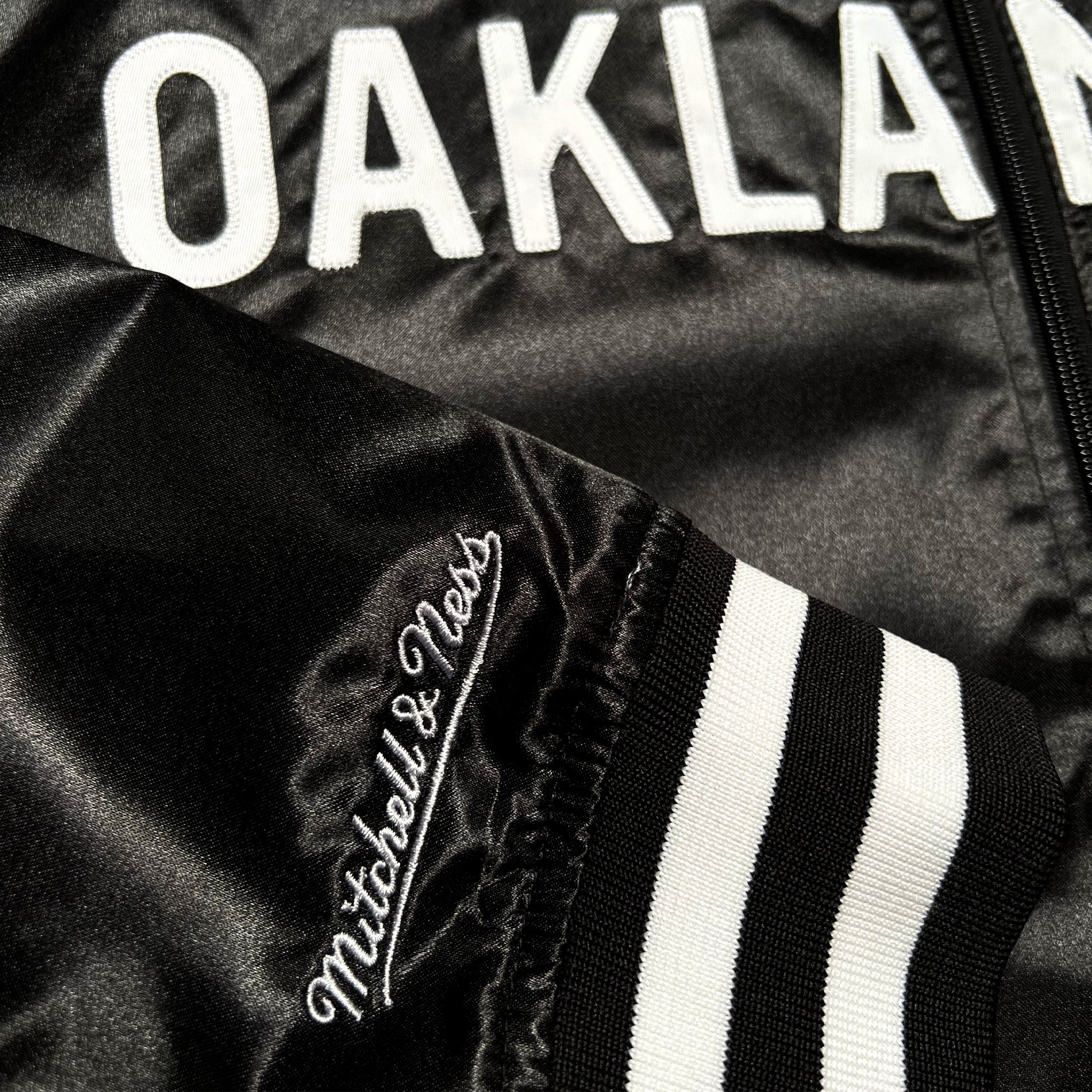 The Mitchell & Ness x Oaklandish Reversible Satin Jacket features "OAKLAND" in bold white across the chest. The sleeve displays white embroidered "Mitchell & Ness" branding with black and white striped ribbed cuffs, offering a smooth, glossy texture.