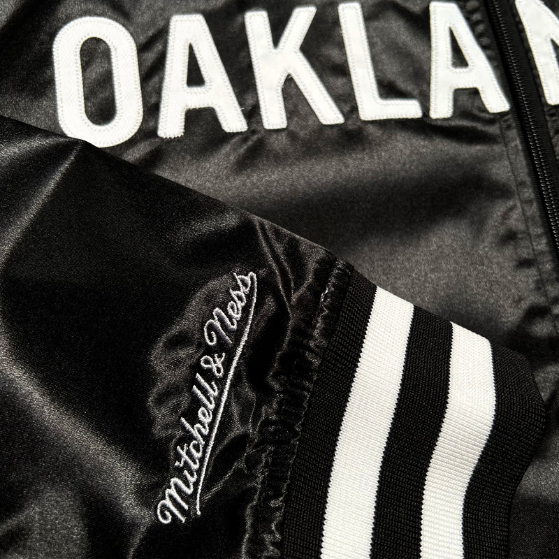Detailed close-up of Mitchell and Ness cursive wordmark on the black and white striped cuff of a black satin jacket.