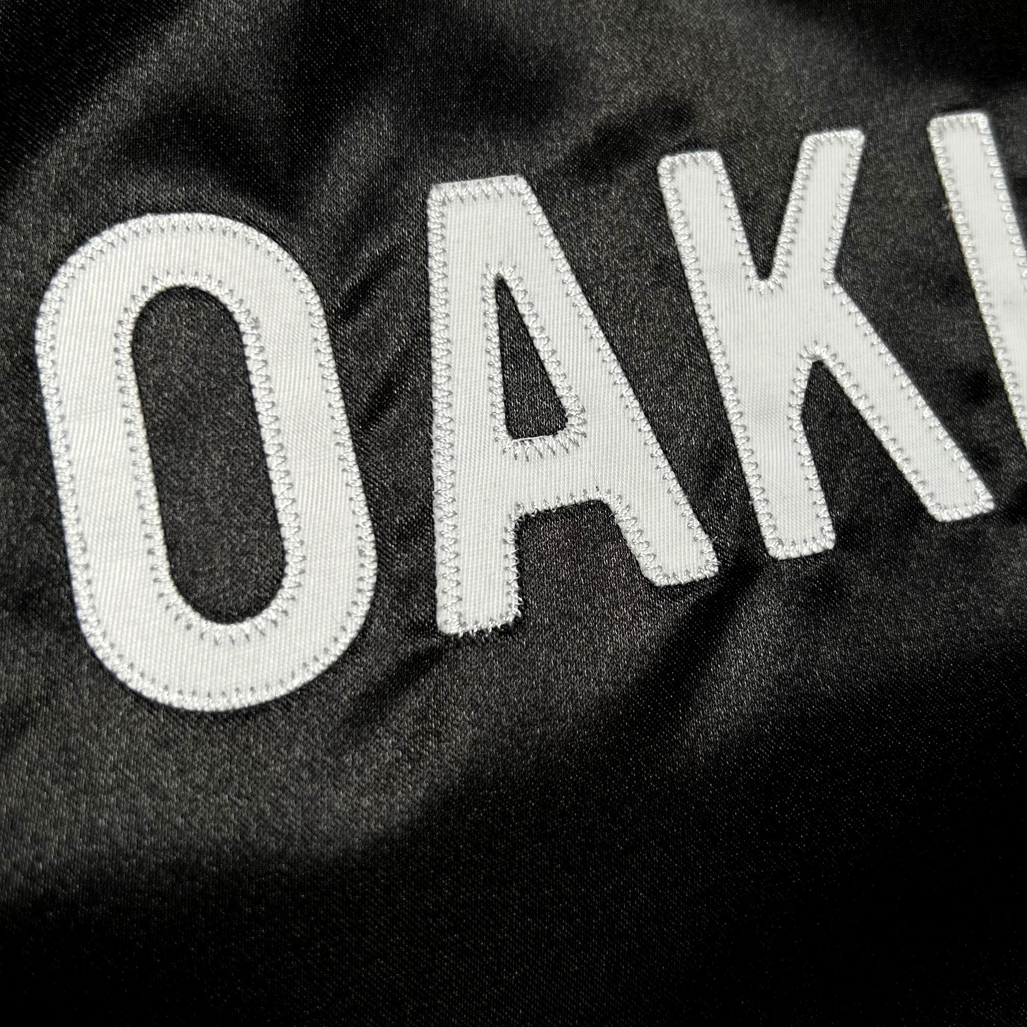 The Mitchell & Ness x Oaklandish Reversible Satin Jacket features "OAKLAND" embroidered in bold white capitals on black fabric. The textured ripples enhance depth, and the prominent stitching highlights the craftsmanship of this Oaklandish apparel by Other.