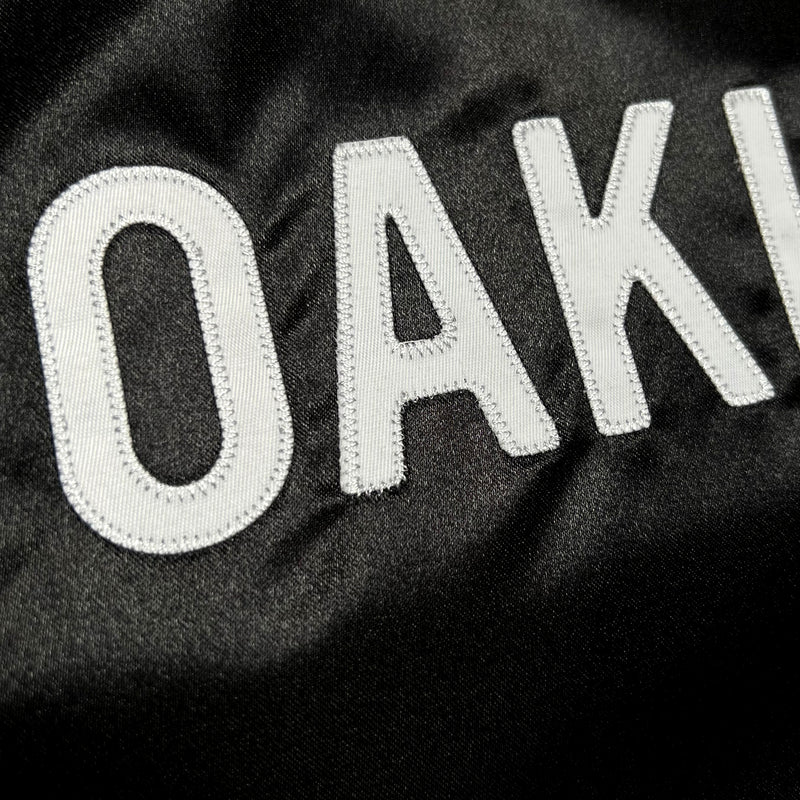 Detailed close-up of OAK letters in Oaklandish wordmark on a black satin reversible jacket.