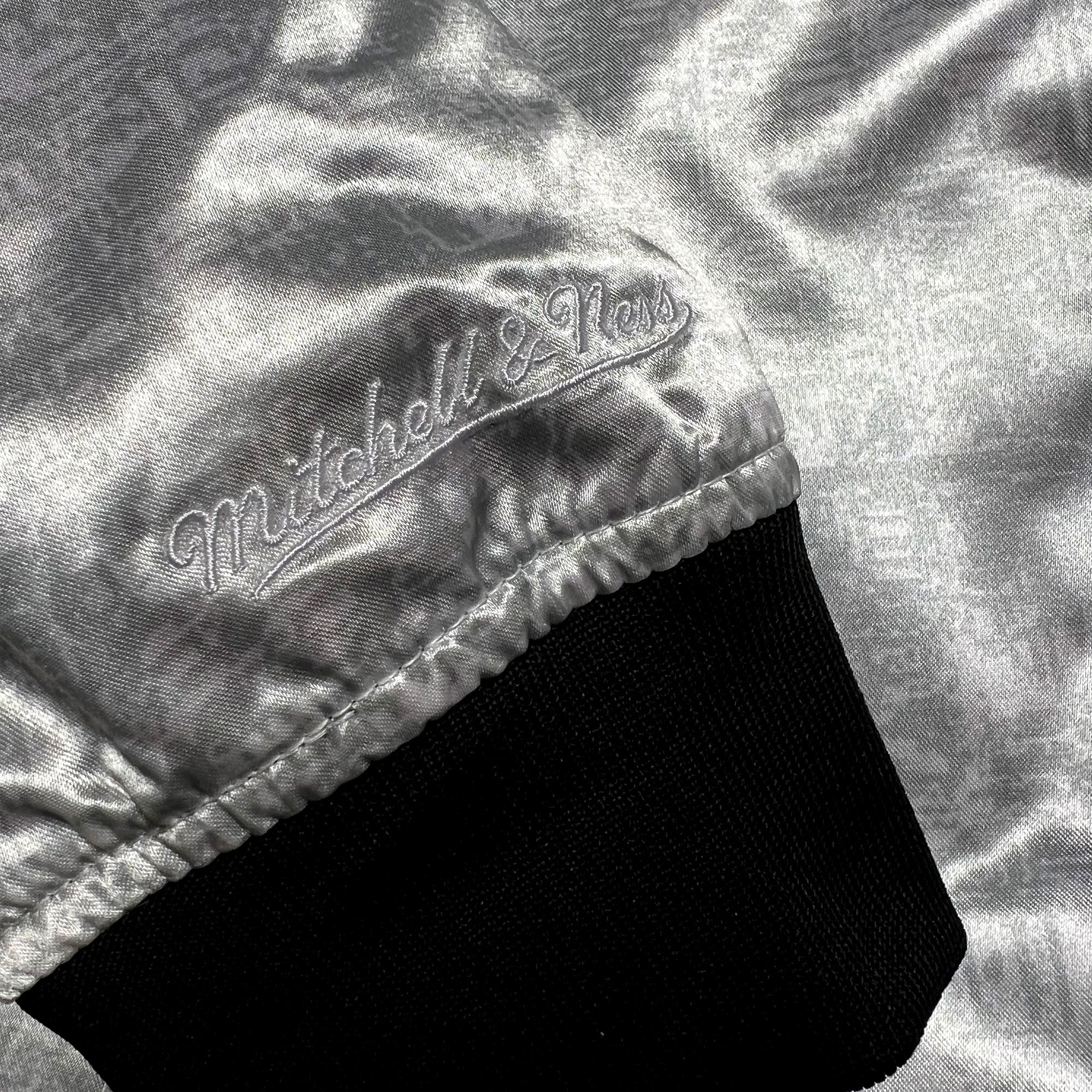 A close-up shows the shiny silver fabric of the Mitchell & Ness x Oaklandish Reversible Satin Jacket, embroidered with "Mitchell & Ness" in cursive. It features a black ribbed cuff with a reflective satin-like texture, enhancing its opulent feel.