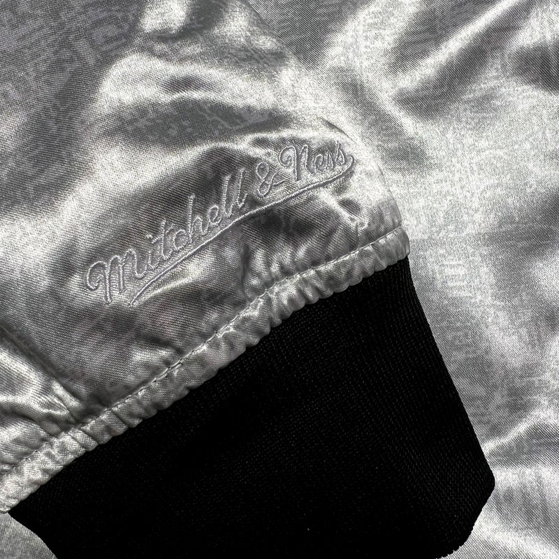 Detailed close-up of Mitchell and Ness cursive wordmark on the black and white striped cuff of a silver reverse side of a satin jacket.