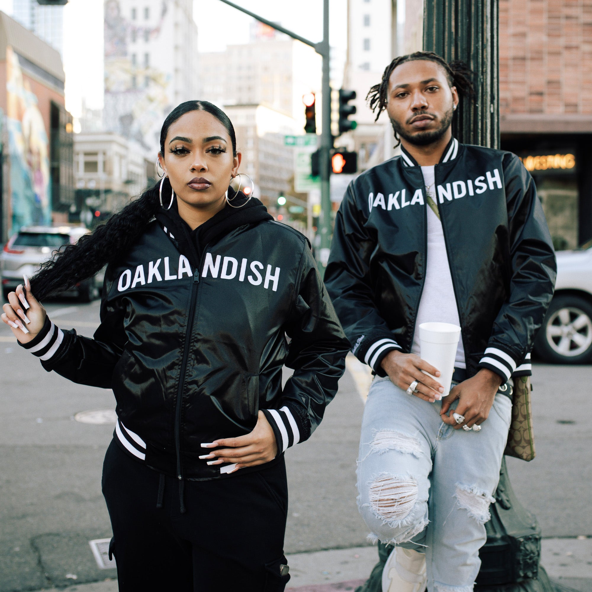 A woman adjusts her ponytail, wearing a Mitchell & Ness x Oaklandish Reversible Satin Jacket alongside a man with ripped jeans and a drink, as they pose on a city street. Urban buildings complete the stylish scene.
