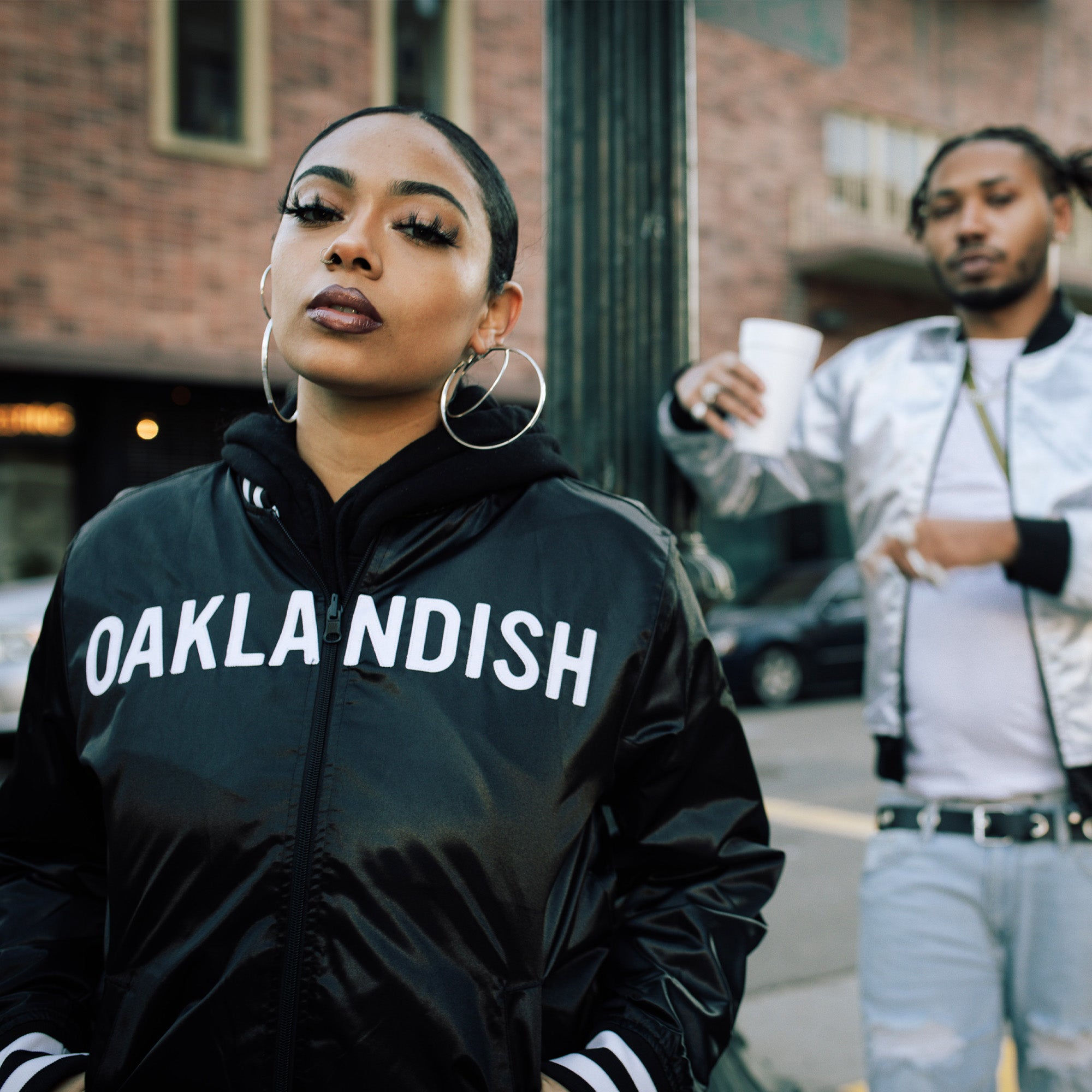 A woman with large hoop earrings and dark lipstick rocks a black jacket in the foreground. Behind her, a man animatedly gestures while holding a cup and wearing the Mitchell & Ness x Oaklandish Reversible Satin Jacket on a bustling city street with brick buildings and cars.