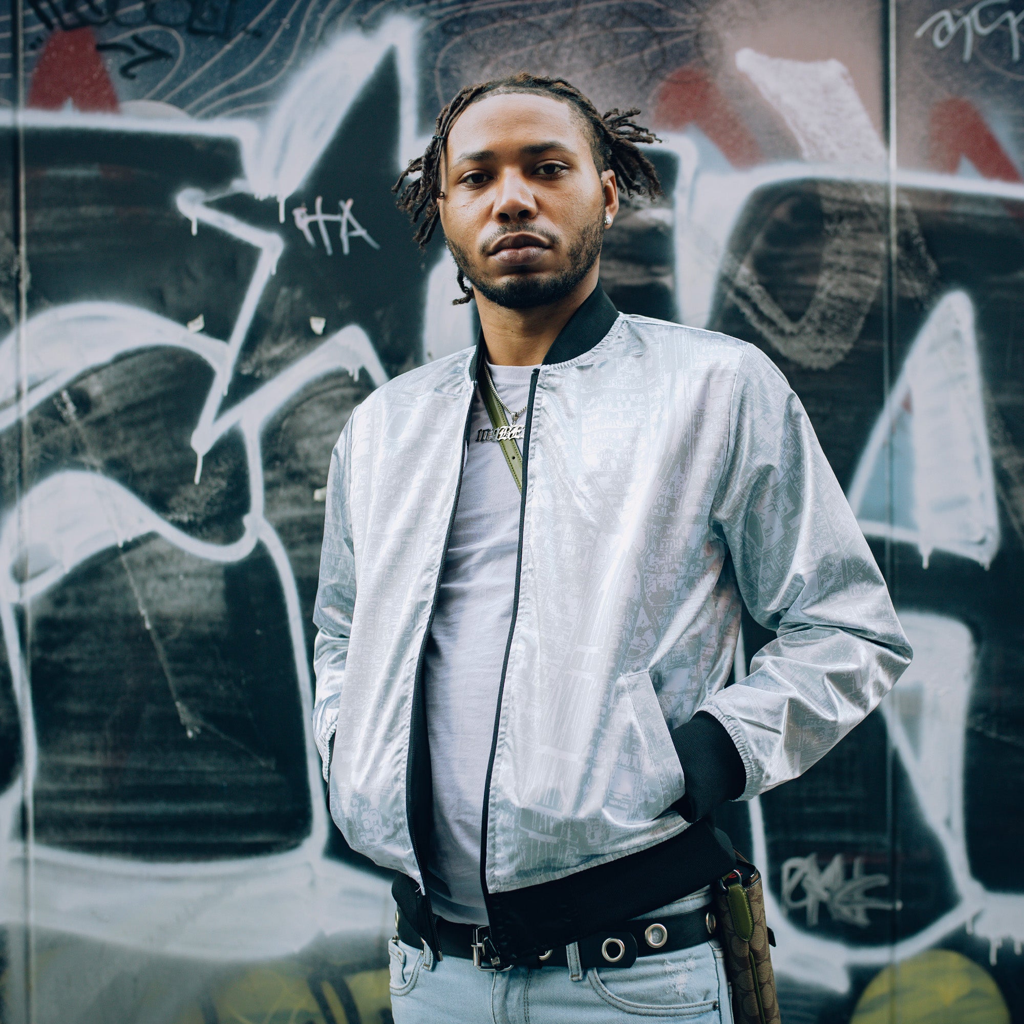 Man outdoors wearing silver reverse side of satin jacket imprinted with an aerial map of Oakland with black ribbing at collar, cuff, and waistband.