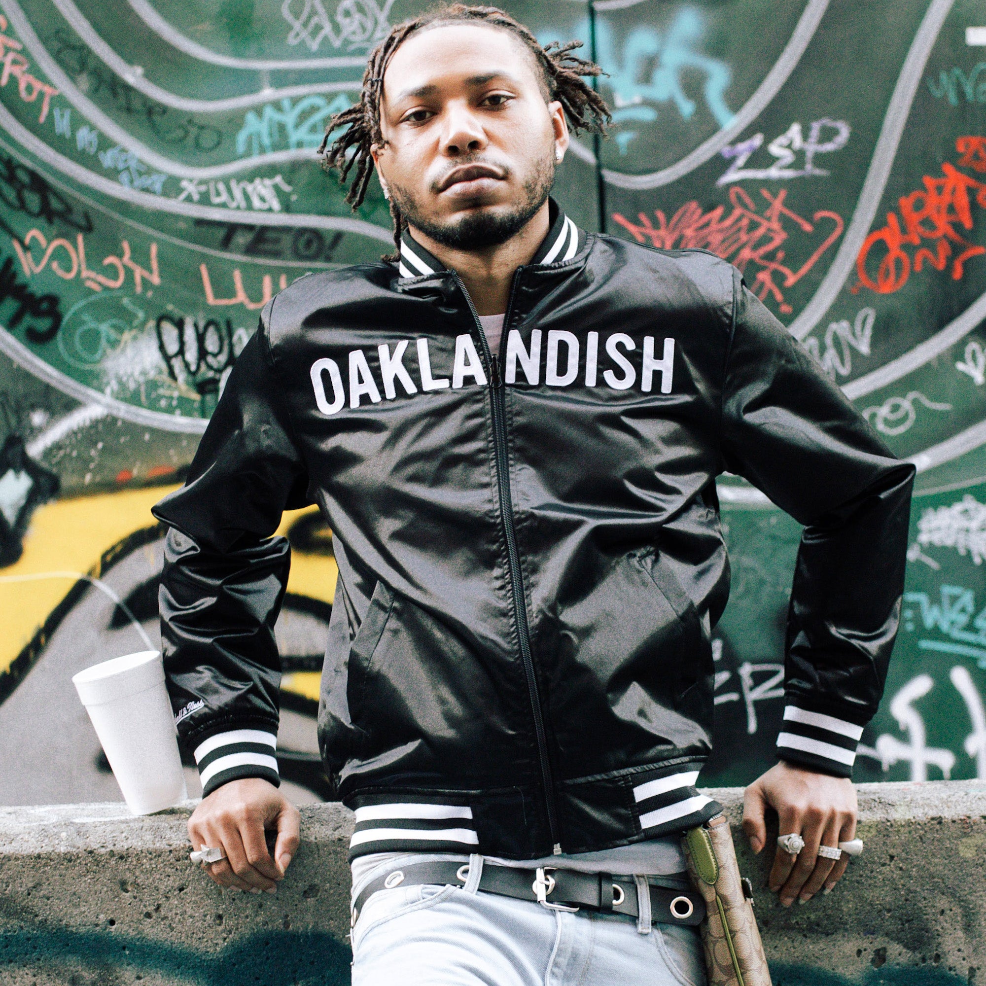A man confidently stands against a graffiti-covered wall, wearing a black Mitchell & Ness x Oaklandish Reversible Satin Jacket with light blue jeans. Braided hair frames his face as rings adorn his hands. A white cup is nearby, and a leather bag strap is visible over his shoulder, completing the look.