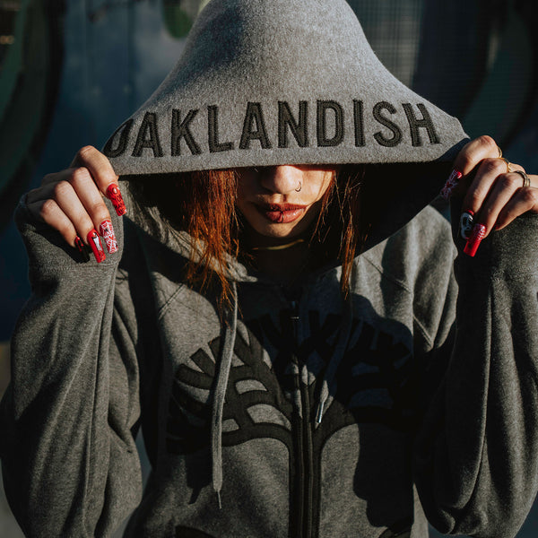 Female model wearing a grey zip-up hoodie with charcoal Oaklandish tree appliqué on the chest and hood up.