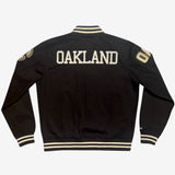Backside of a black zip-up cadet jacket with Oaklandish wordmark across the back and black and white striped ribbing on the bottom, the cuffs & the neck.