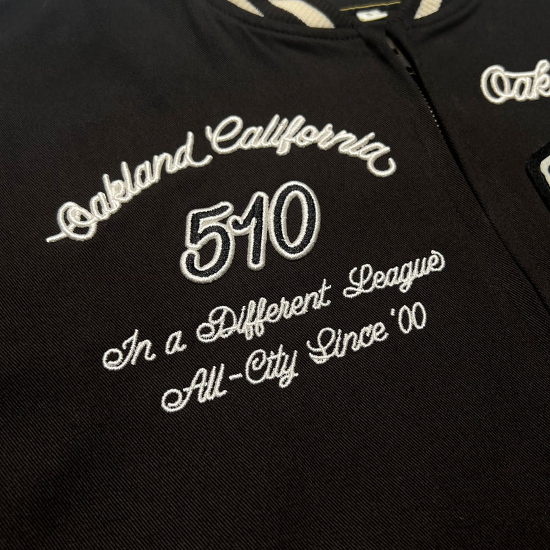 Close-up of cursive embroidery on the chest of a black Oaklandish cadet jacket saying “Oakland California 510 in a different league all-city since '00'.