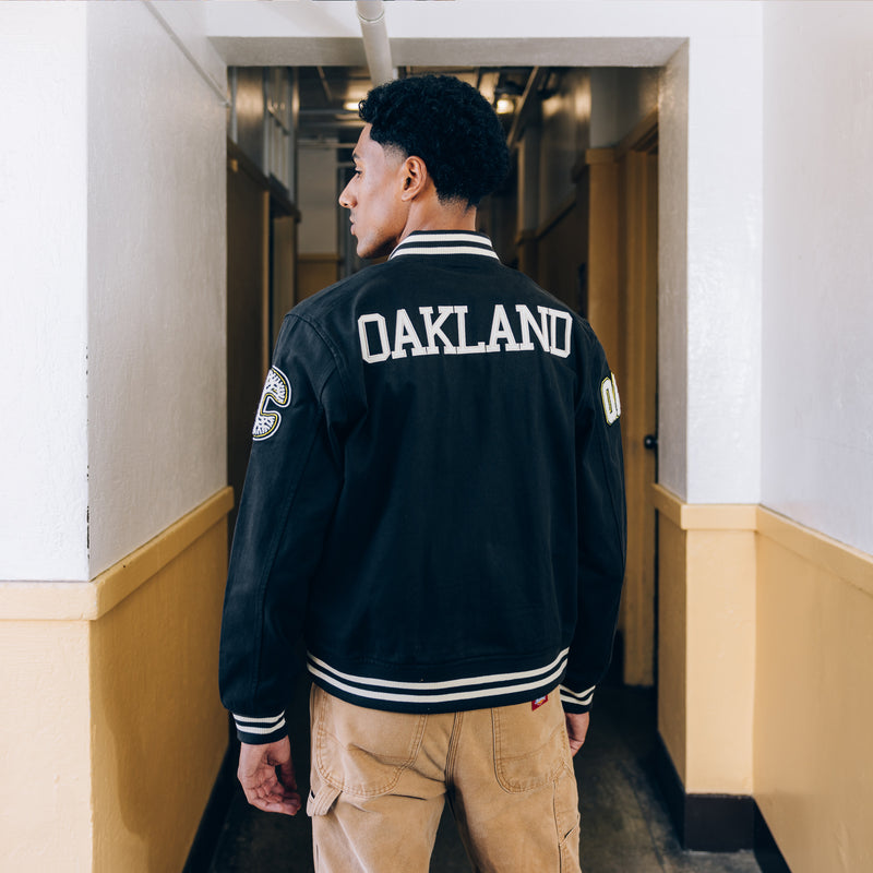 A man standing in school, back to the camera in a black cadet jacket with OAKLAND wordmark on the back and striped ribbing on the bottom and cuffs.