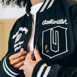 Close-up of a woman’s chest in a black zip-up cadet jacket with Oaklandish chenille patches and striped ribbing.