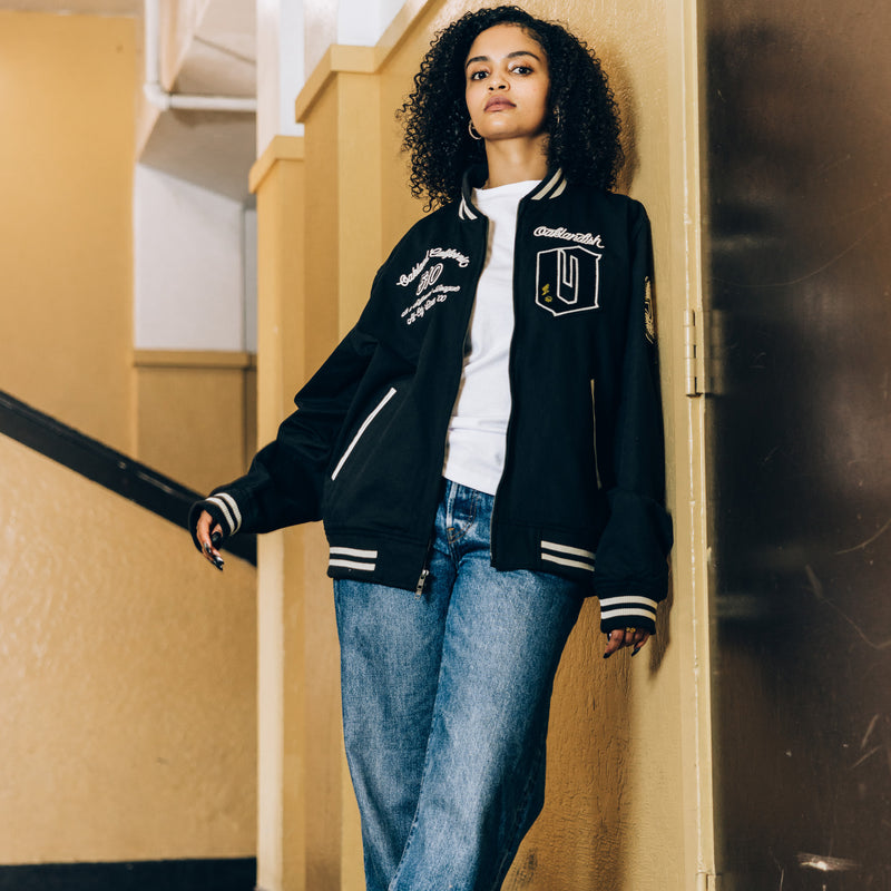 A woman standing in a school in a black cadet jacket with Oaklandish chenille patches and black and white striped ribbing.