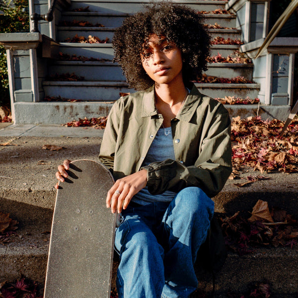 Woman sitting on an outdoor stoop holding a skateboard wearing an olive greencoaches jacket with an 'Oaklandish' wordmark on the left chest.