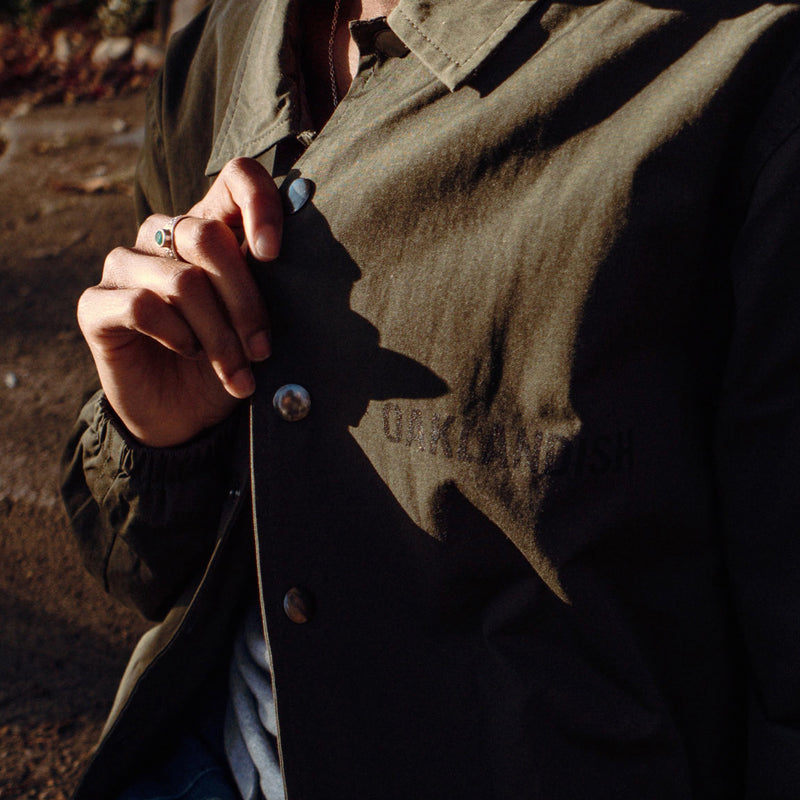 Close-up of the black Oaklandish wordmark on the left chest of an olive green coaches jacket worn by a woman standing outdoors.