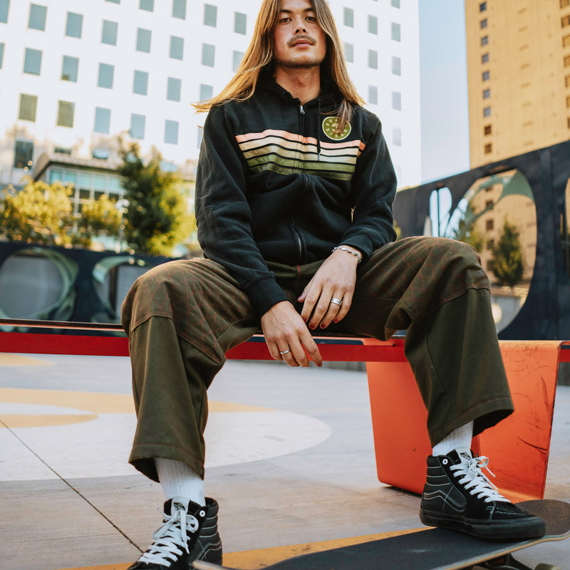 A male model sitting on a bench with skateboard wearing a black Oaklandish zip-up hoodie.