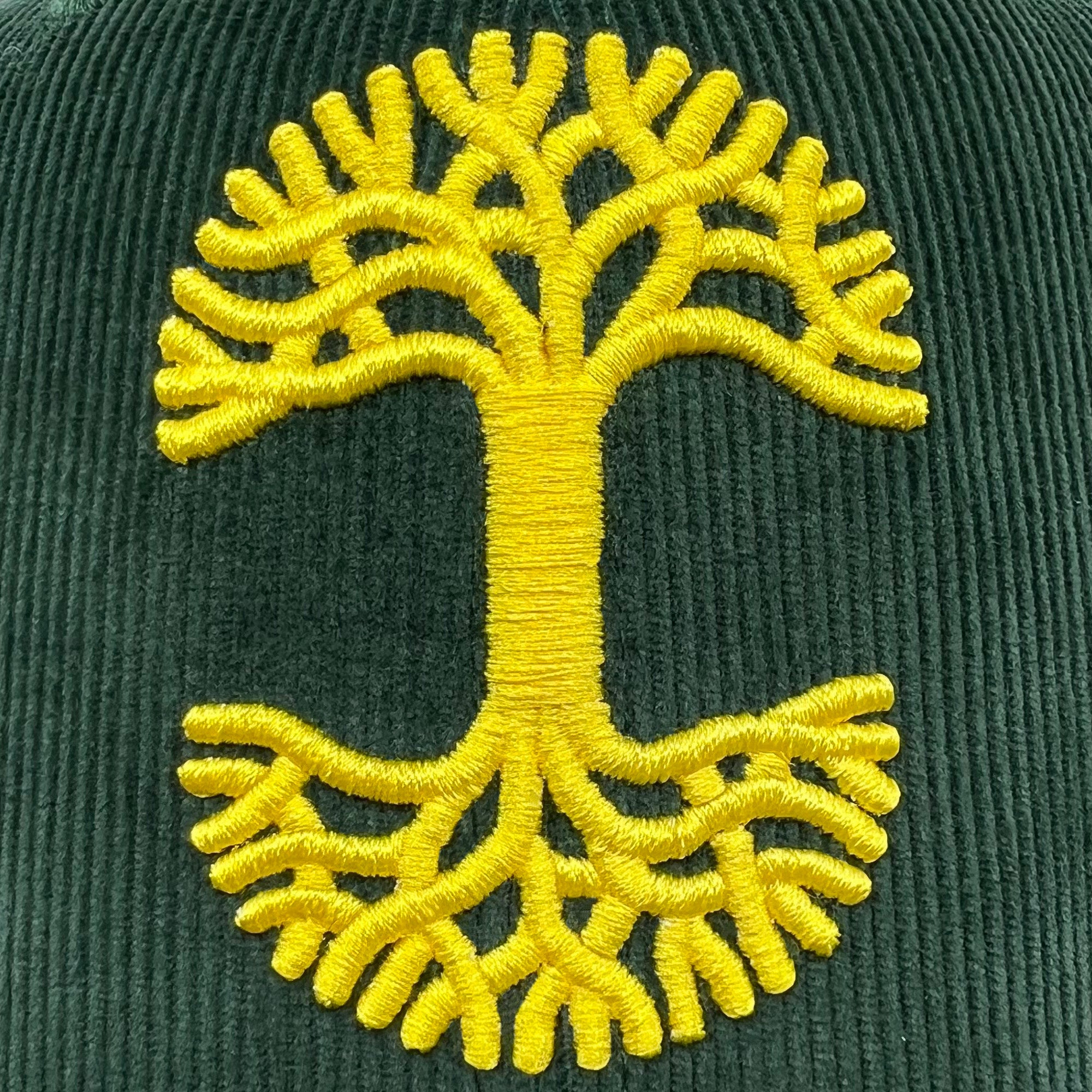 Detailed close-up of embroidered yellow Oaklandish tree logo on a forest green corduroy Mitchell & Ness cap.