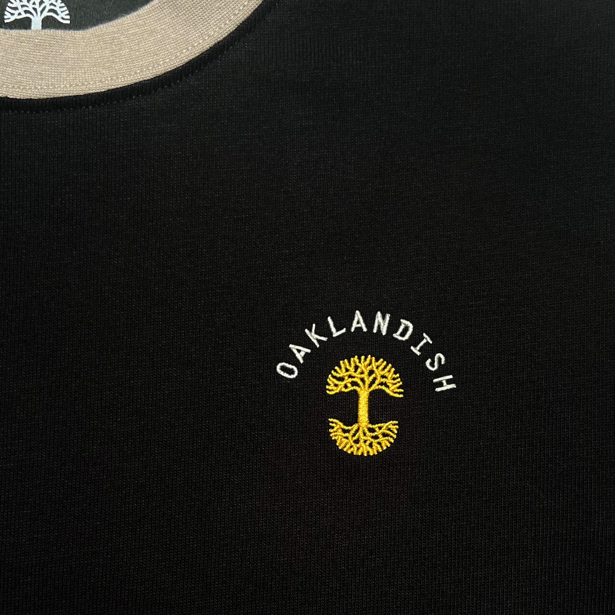 Close-up of embroidered gold Oaklandish tree logo, white wordmark on the chest of a black long-sleeve t-shirt.