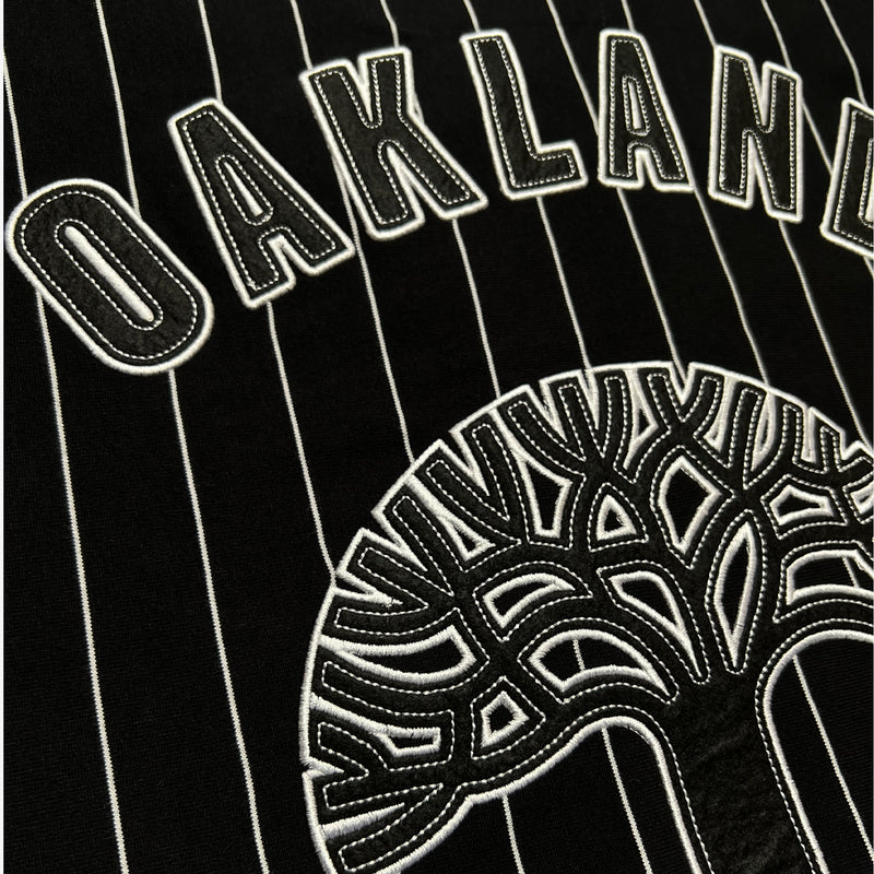 Detailed close-up of large black and white Oaklandish tree logo and wordmark applique on the back of black baseball jersey.