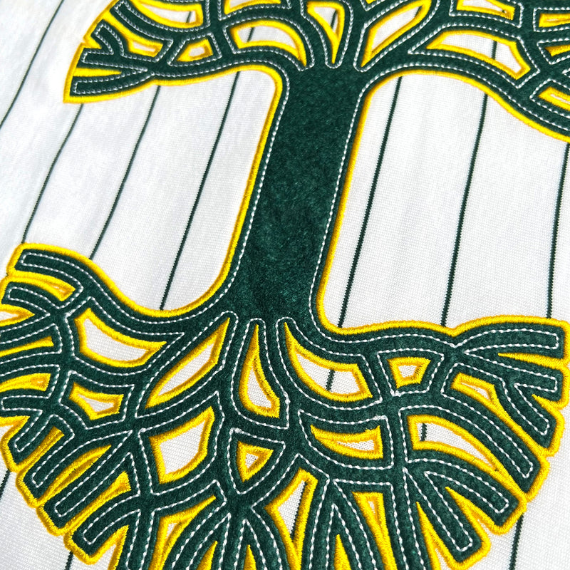 Detailed close-up of a large green and yellow Oaklandish tree logo on a white baseball jersey.