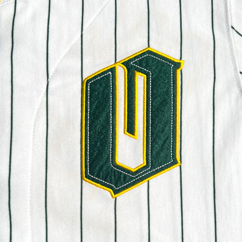 Detailed close-up of green and gold O for Oakland applique on the chest on a white baseball jersey.