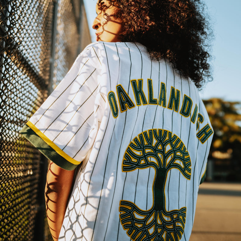 Close-up of a large green and yellow Oaklandish tree logo and wordmark applique on a white baseball jersey on a woman on a baseball field.